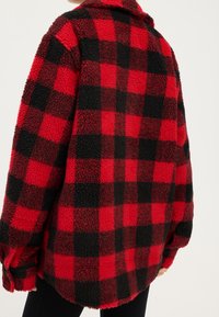 Rød og sort ternet fleece-skjortejakke med en tekstureret, fluffy overflade, lange ærmer og en løs pasform, med krave.