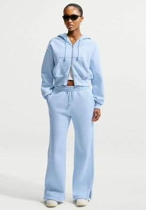 Kvinde iført lyseblå hættetrøje med lynlås og matchende vidde sweatpants, hvide sneakers og sorte solbriller, stående foran en ensfarvet baggrund.