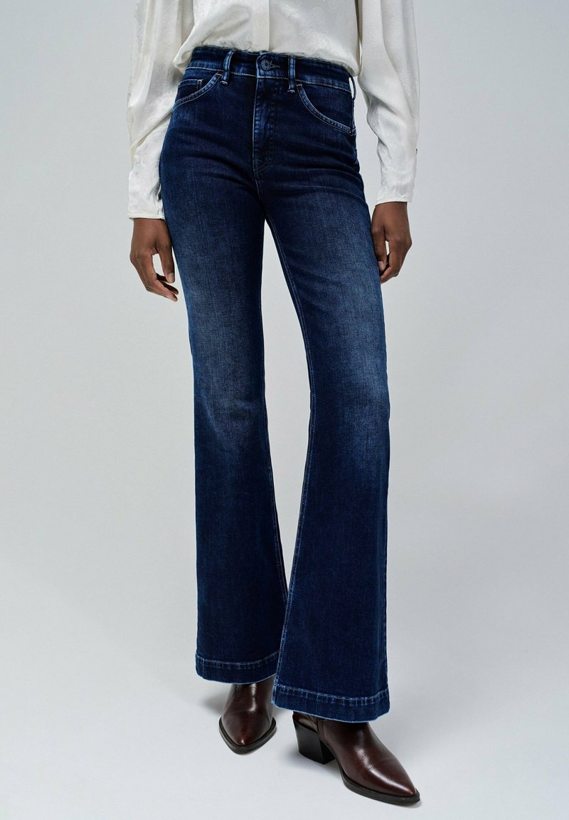 Femme portant un jean évasé taille haute bleu foncé, un chemisier blanc à manches longues et des bottines marron foncé à bouts pointus sur fond gris.