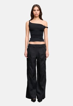 Femme debout, face à nous, portant un haut noir asymétrique sans manches, un pantalon noir ample et des chaussures noires pointues sur un fond uni.