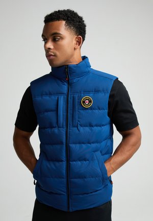 Mann trägt eine blaue gesteppte ärmellose Weste mit Zavetti Canada-Logo, ein schwarzes kurzärmeliges Shirt, steht mit den Händen in den Taschen und schaut nach links.