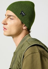 Berretto lavorato a coste verde con etichetta nera Wrangler; indossato con un gilet isolante verde oliva e una maglietta a maniche lunghe abbinata.