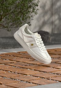 Λευκό δερμάτινο αθλητικό παπούτσι με λείο φινίρισμα, σχέδιο με κορδόνια, διακριτικές επενδύσεις με μαξιλαράκια και χρυσό έμβλημα στο πλάι.
