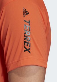 Orange atletisk t-shirt med korte ærmer; har sort "adidas" logo og "TERREX" tekst i fed skrift på ærmet; glat stof tekstur.