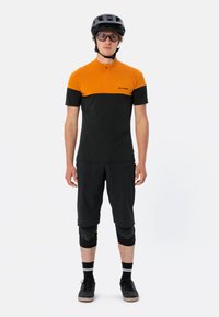 Maillot de vélo à manches courtes avec des blocs de couleur orange et noir, col zippé et logo. Associé avec un short noir et des genouillères.