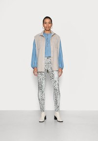 Colete bege sem mangas sobre uma camisa azul de botão, combinado com leggings de padrão de cobra e botas brancas de tornozelo com salto quadrado.