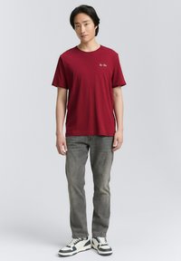 Rød bomulds T-shirt med korte ærmer og rund hals, parret med grå jeans og sort-hvide sneakers. Minimalistisk logo detalje.