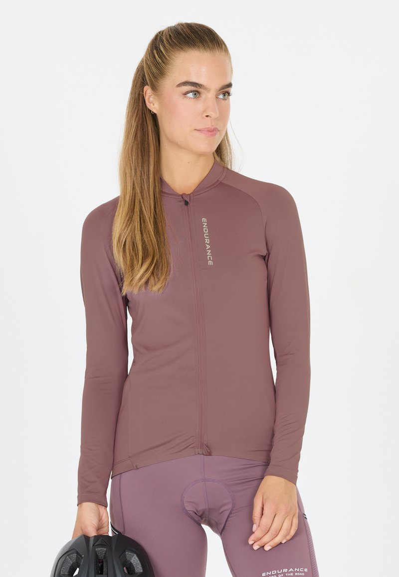 Vrouw met lang haar, gekleed in een mauve wielershirt en korte broek, houdt een zwarte fietshelm vast en kijkt opzij tegen een witte achtergrond.