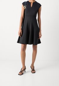 Robe noire à manches courtes style casquette, avec un décolleté en forme de goutte, un corsage ajusté et une jupe évasée. Associée à des sandales noires à talons hauts et lanières.