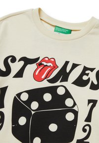 Cremefarbendes Baumwoll-T-Shirt mit einem großen roten Zungenlogo, fettem schwarzen Schriftzug und einer Grafik eines schwarzen Würfels mit weißen Punkten.