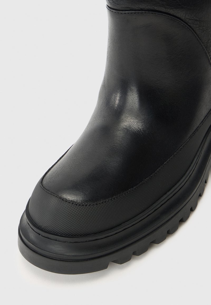 Sort støvle med en glat læderoverdel, tekstureret gummisål og rund tå. Bemærkelsesværdig for sine kontrasterende materialer og elegante design.