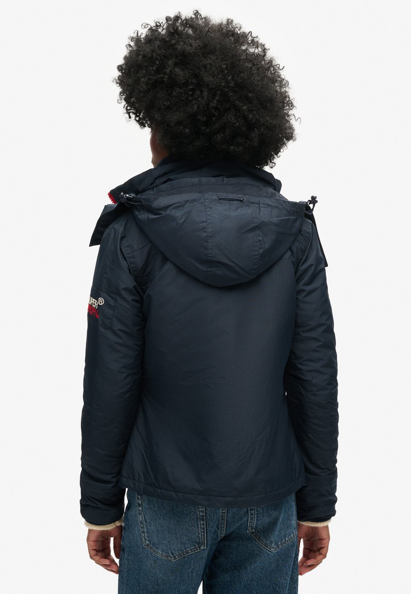 Persoon met krullend haar, gekleed in een navy blauwe capuchonjack en blauwe jeans, staat met de rug naar de camera tegen een witte achtergrond.