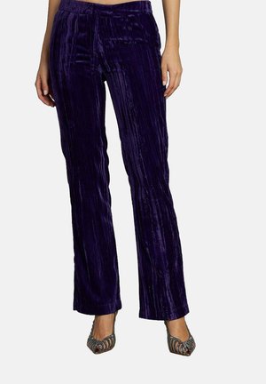 Pantalons en velours violet avec une coupe droite, ornés de côtes verticales. Portés avec des escarpins ouverts scintillants.