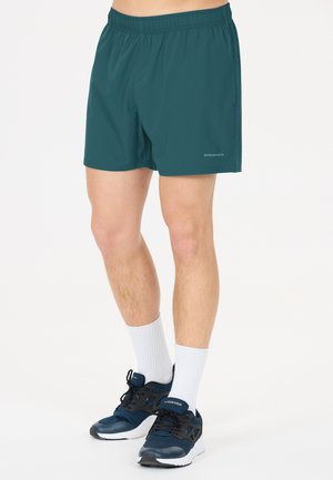 Mandeben iført grønne træningsshorts, hvide mellemlange strømper og mørkeblå løbesko på en ensfarvet hvid baggrund.