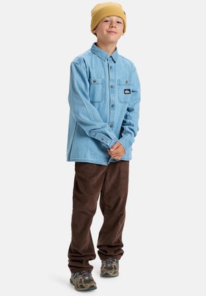 Jongen met een gele muts, een lichtblauw knoopshirt, een bruine corduroy broek en grijze sneakers, staat met de handen gevouwen.