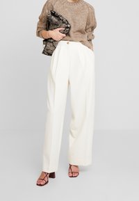 Beige neulepaita, valkoiset levenevät housut pliseerauksilla ja remmikkäät korkokengät. Pitelee mustavalkoista, teksturoitua laukkua.