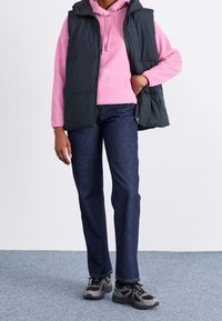 Persoon in een roze hoodie, zwart gewatteerd vest, donkerblauwe jeans en zwart-grijze sneakers die op een blauwe tapijtvloer staat.