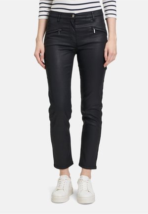 Černé slim-fit kalhoty vyrobené z elastického materiálu, s kapsami na zip a hladkým povrchem. Nosí se s bílými teniskami.