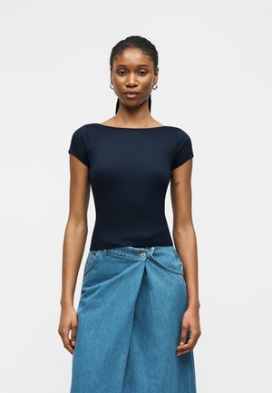 Femme aux cheveux tressés portant un haut bleu marine ajusté à manches courtes et une jupe en jean bleue asymétrique, debout devant un fond uni.