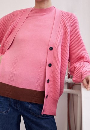 Personne portant un cardigan en maille rose avec des boutons noirs par-dessus un pull rose et marron, associé à un jean bleu, debout à l'intérieur.