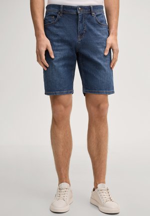 Mężczyzna ubrany w niebieskie dżinsowe szorty do połowy uda i białe trampki, stojący na tle gładkiego tła.