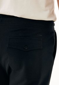 Černé kalhoty s zadní kapsou na knoflíkový uzávěr. Tkanina je hladká s mírným leskem a logo je umístěno nad kapsou.