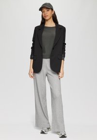 Schwarzes Blazer mit Knopfverschlüssen, grauer Strickoberteil, karierte weite Hose, graue Mütze und silberne Sportschuhe. Weiche Texturen und taillierter Schnitt.