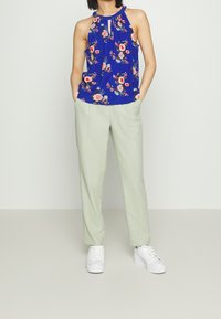 Modrý květinový top bez rukávů s výstřihem ve tvaru klíče, spárovaný se světle zelenými kalhotami a bílými teniskami. Hladké tkaniny a uvolněný střih.