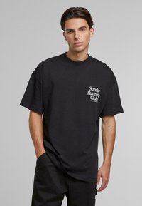Black cotton t-shirt with oversized fit, crew neck, and left chest graphic reading "Sunday Running Club" in white lettering.