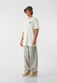 Tricou alb din bumbac cu logo negru, croială lejeră, purtat cu blugi gri deschis, largi, și adidași bej cu un motiv floral. Pălărie neagră tip bucket.