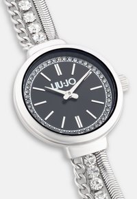 Silberne Metalluhr mit schwarzem Zifferblatt, Kristallakzenten und einem Gliederarmband. Verfügt über ein rundes Gehäuse und silberne Stundenmarkierungen.