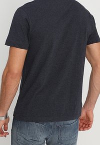 Tmavě šedé tričko s krátkým rukávem vyrobené z měkkého materiálu. Má klasický kulatý výstřih a jednobarevný zadní design. Nosené s vybledlými modrými džíny.