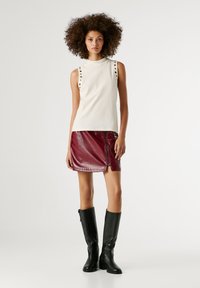 Fiatal nő göndör hajjal, ujjatlan, szegecses krém színű felsőt visel, fényes burgundi mini szoknyát és térdig érő fekete csizmát.