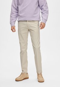 Chino beige avec une texture lisse, coupe slim et poches latérales, associés à un sweatshirt violet clair et des chaussures en daim beige.
