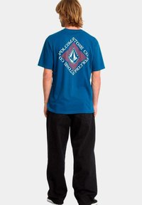 Hombre con cabello rubio vistiendo una camiseta azul de Volcom con un gran logo geométrico y texto en la parte de atrás, pantalones negros y zapatillas negras, de espaldas.