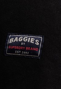 Etiqueta de tela negra con el texto "BAGGIES by SUPERDRY BRAND EST 2003" en blanco y rojo, sobre un fondo azul marino y con costuras blancas.