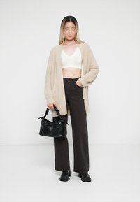 Beige, fluffy cardigan over een witte crop top, gecombineerd met donkerbruine wijde broekspijpen. Houdt een kleine zwarte handtas met een gestructureerde afwerking vast.
