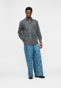 Άνδρας που φοράει μακρυμάνικο πουκάμισο με μαύρες και λευκές κάθετες ρίγες, παντελόνι φαρδύ με μπλε σχέδιο και μαύρα παπούτσια, στέκεται μπροστά σε λευκό φόντο.