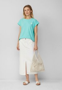 Vrouw in een turquoise mouwloos top en een witte midi rok, met een beige netten tote bag, en witte instap sandalen.