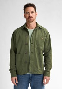 Green button-up jacket made of cotton, featuring a chest pocket, two front pockets, and a collar, paired with a light green shirt underneath.