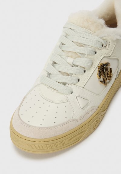 White leather sneaker featuring a fluffy interior, round toe, perforated detailing, and a gold emblem on the side, with a beige suede toe cap.