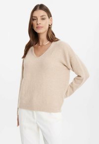 Maglione beige a collo a V realizzato in morbido tessuto testurizzato. Presenta spalle scese e polsini a coste, abbinato a pantaloni bianchi.