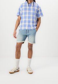 Koszula w kratkę z krótkim rękawem w jasnoniebieskich i fioletowych odcieniach, zestawiona z jasnoniebieskimi szortami dżinsowymi, białymi skarpetkami oraz beżowymi trampkami z ciemnymi akcentami.