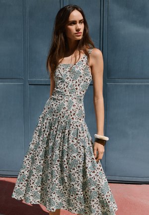 Moteris dėvi be rankovių, raštuotą midi suknelę su žaliomis ir rudomis gėlių detalėmis, stovinti prieš mėlyną skydinę sieną.