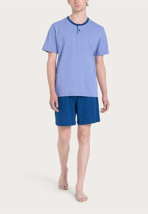Uomo che indossa una camicia a maniche corte azzurro chiaro con colletto blu scuro e pantaloncini blu scuro abbinati, in piedi a piedi nudi su sfondo bianco.