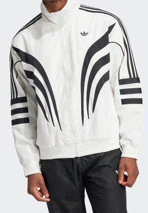 Homme portant une veste blanche zippée avec des éléments de design noirs courbés et rayés et un pantalon noir avec un logo sur la cuisse gauche.