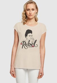 Beige T-Shirt mit kurzen, aufgerollten Ärmeln, versehen mit einer schwarzen Linzeichnung eines Frauenkopfes und dem Wort "Rebel" in einer eleganten Schriftart, akzentuiert mit Blumen.