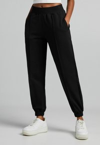 Svarta joggerbyxor med smockad midja, sidofickor och elastiska muddar. Slätt material med avslappnad passform. Kombinerade med vita sneakers.