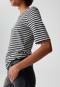 Personne portant une chemise à rayures noir et blanc à manches courtes rentrée dans un jean foncé, vue de côté taille haute contre un fond gris.