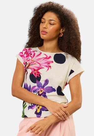 Naiste lokkis juustega, kes kannab beežikat lühikeste varrukatega pluusi suurte mustade täppide ning roosade ja lilla lillemustritega, mida kannab heledate roosade pükstega.